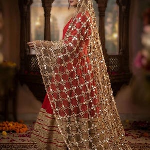 Puede incluir: Un vestido rojo con adornos dorados y un velo transparente de color oro. El velo está adornado con pequeños adornos reflectantes. El vestido parece estar hecho de una tela fluida, lo que sugiere una prenda formal o cultural.