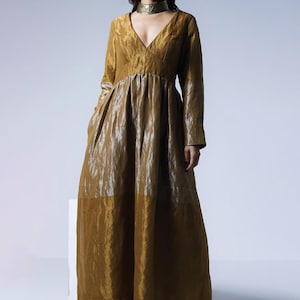 May include: A long, flowing gold dress with a V-neck and long sleeves. The dress has a gathered waist and a wide skirt. The model is wearing gold heels and a gold choker. The dress is made of a shiny, textured fabric.