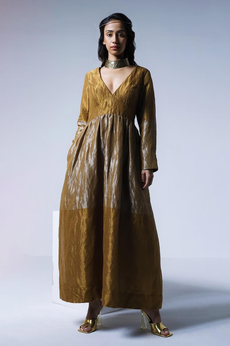 May include: A long, flowing gold dress with a V-neck and long sleeves. The dress has a gathered waist and a wide skirt. The model is wearing gold heels and a gold choker. The dress is made of a shiny, textured fabric.