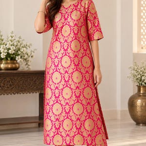 May include: A bright pink knee-length tunic with three-quarter sleeves and matching straight-leg trousers, both featuring a repeating gold floral brocade pattern. The tunic has a round neckline and side slits, paired with gold-toned flat shoes.