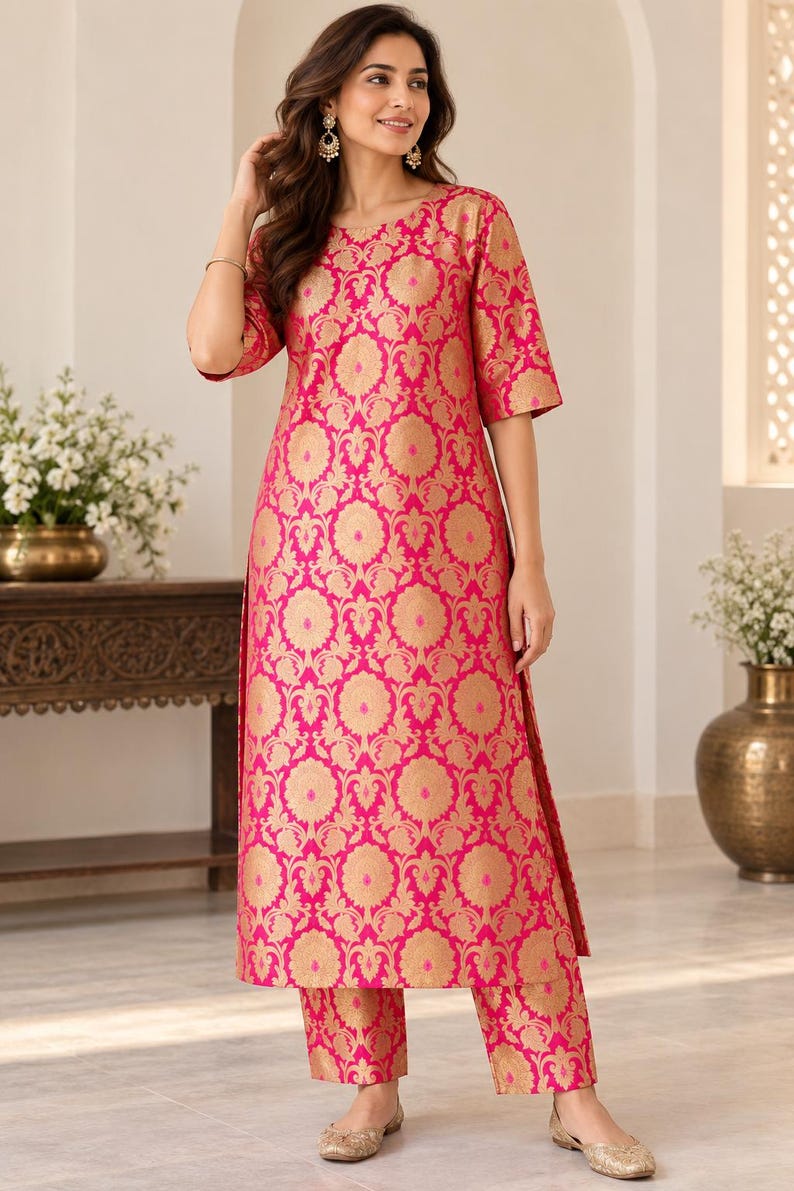 May include: A bright pink knee-length tunic with three-quarter sleeves and matching straight-leg trousers, both featuring a repeating gold floral brocade pattern. The tunic has a round neckline and side slits, paired with gold-toned flat shoes.
