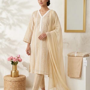 May include: A beige embroidered salwar kameez outfit. The ensemble features a long-sleeved tunic with a V-neck, matching trousers, and a sheer scarf. The tunic and trousers are decorated with white floral embroidery. The outfit is paired with beige flats.