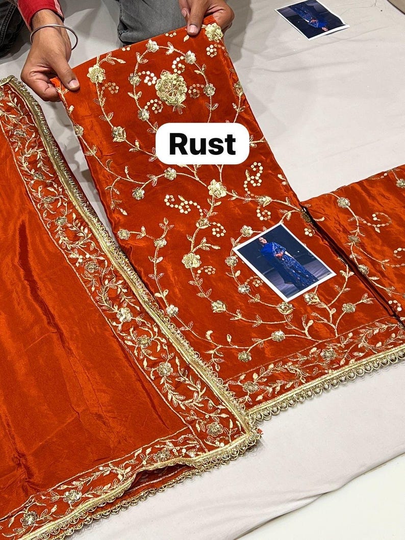 May include: Rust-coloured fabric with detailed gold embroidery, showcasing floral and vine designs. The fabric is presented with a matching border and a small image. The word "Rust" is displayed on the fabric.