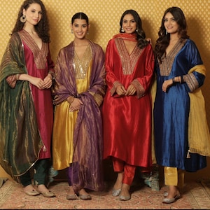 May include: Four women wearing traditional Indian clothing. The women are wearing long, flowing dresses with intricate embroidery and colorful dupattas. The dresses are in shades of red, blue, gold, and green. The women are standing in front of a gold background.