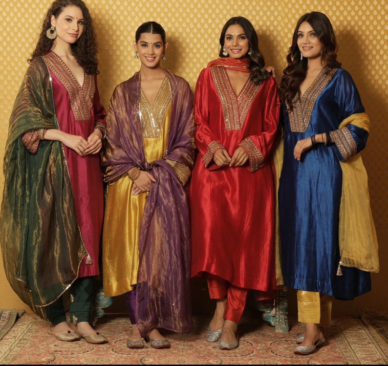 May include: Four women wearing traditional Indian clothing. The women are wearing long, flowing dresses with intricate embroidery and colorful dupattas. The dresses are in shades of red, blue, gold, and green. The women are standing in front of a gold background.