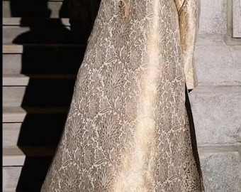 Pode incluir: Uma túnica dourada de manga comprida com um padrão floral de brocado intrincado, gola alta com carcela de botões central e um detalhe de bainha em verde-azulado escuro. A peça é combinada com calças estampadas a condizer e brincos dourados.