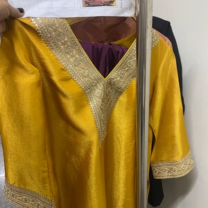 May include: A mustard yellow tunic with gold embroidery around the neckline and sleeves. The tunic has a V-neckline and is made of a silky fabric.