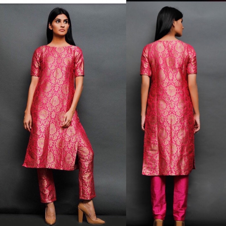 May include: A vibrant fuchsia and gold brocade tunic set featuring a knee-length tunic with short sleeves and a round neckline, paired with matching straight-leg trousers. The fabric displays an intricate floral pattern throughout the garment.