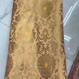 May include: A folded piece of peach-colored fabric featuring an intricate woven pattern in metallic gold thread. The design consists of repeating floral and botanical motifs with small, bright yellow accents centered within the larger blossoms.