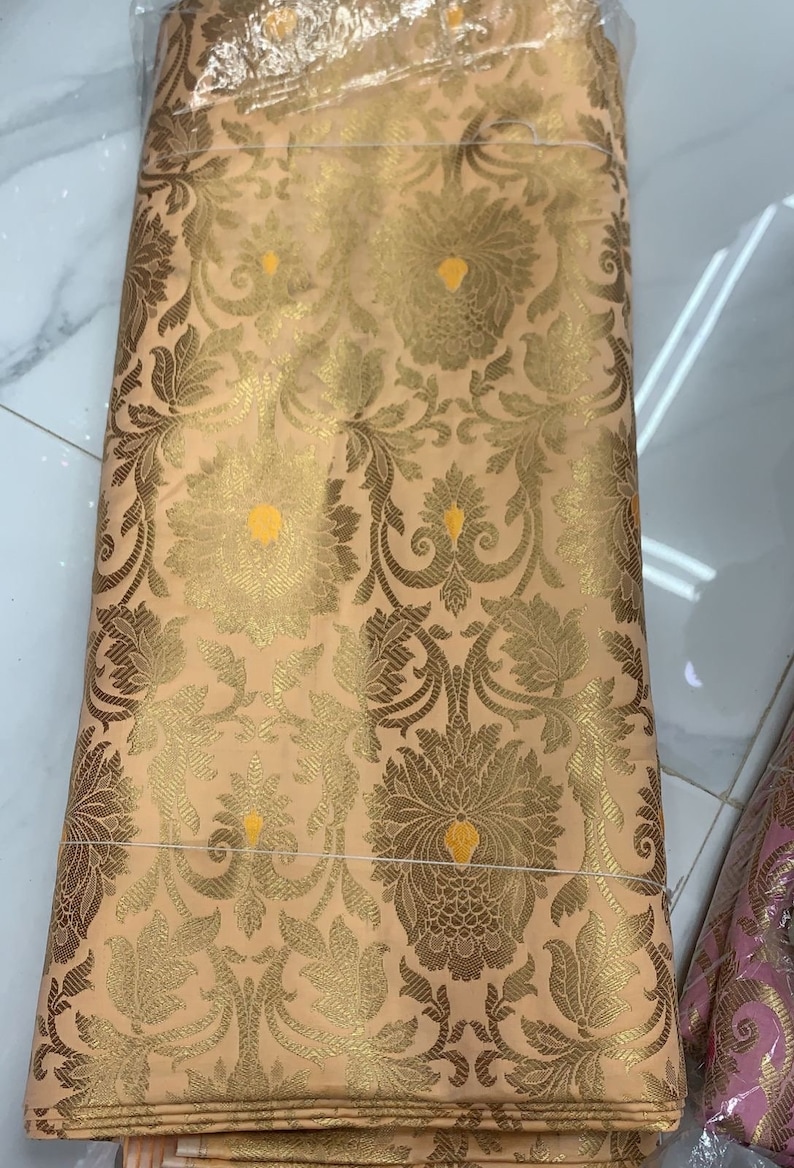 May include: A folded piece of peach-colored fabric featuring an intricate woven pattern in metallic gold thread. The design consists of repeating floral and botanical motifs with small, bright yellow accents centered within the larger blossoms.