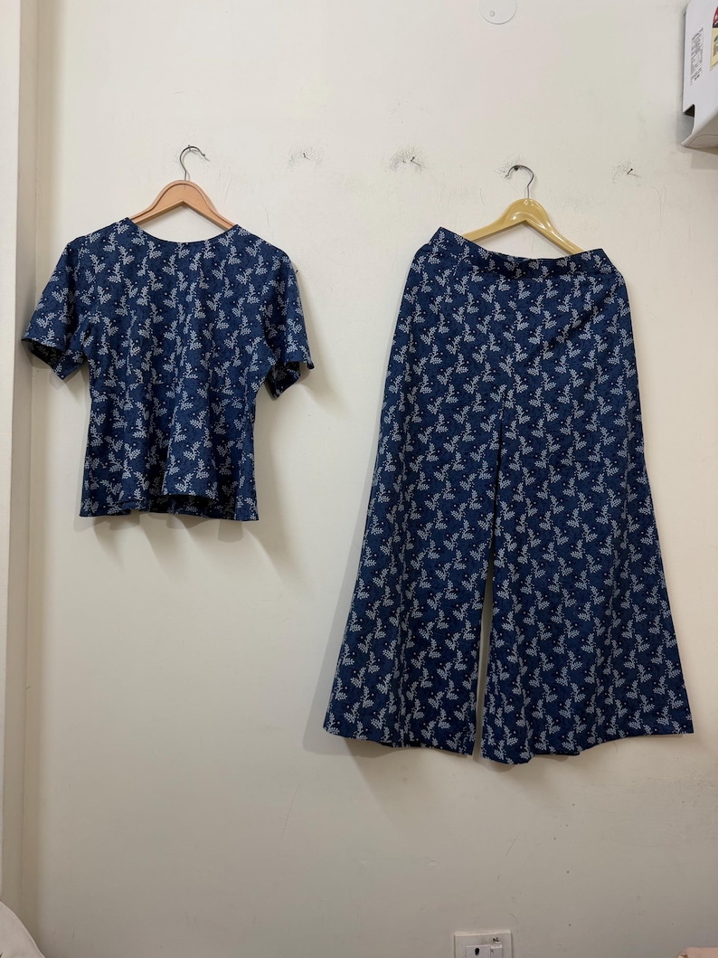 May include: A two-piece set consisting of a short-sleeved top and wide-leg trousers. Both garments are made from a dark blue fabric featuring a repeating white botanical leaf pattern. The top has a round neckline, and the trousers have a high waist.