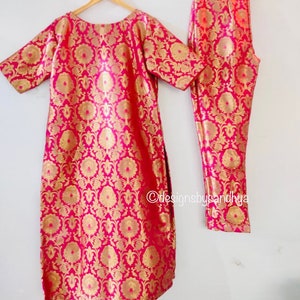 May include: A bright pink and gold metallic brocade tunic and matching trousers set featuring a repeating floral pattern. The tunic has short sleeves and a round neckline. The text designsbysandhya is visible on the lower right side.