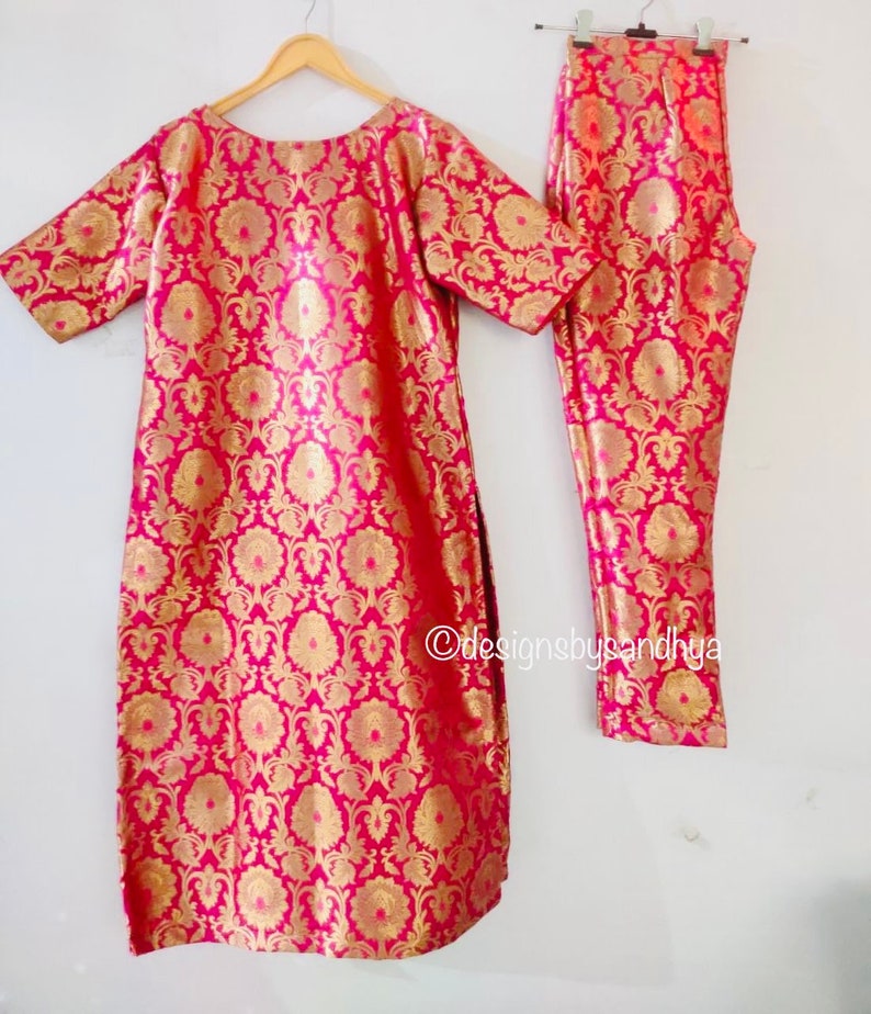 May include: A bright pink and gold metallic brocade tunic and matching trousers set featuring a repeating floral pattern. The tunic has short sleeves and a round neckline. The text designsbysandhya is visible on the lower right side.