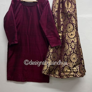 May include: A maroon long-sleeved top with a round neckline and a gold and maroon brocade skirt with a floral pattern. The skirt has a flared bottom. The text "@designsbysandhya" is visible in the image.
