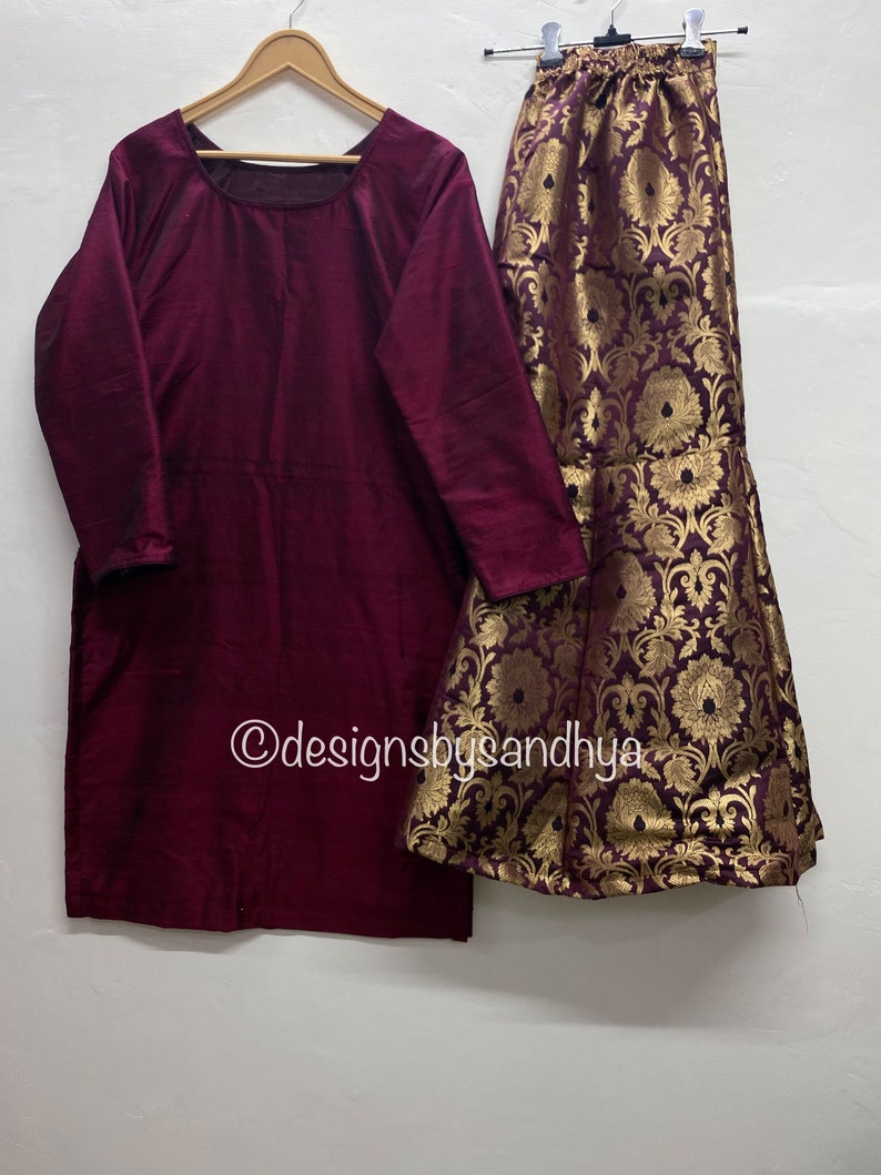 May include: A maroon long-sleeved top with a round neckline and a gold and maroon brocade skirt with a floral pattern. The skirt has a flared bottom. The text "@designsbysandhya" is visible in the image.