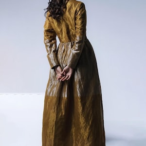 May include: A long, golden-brown dress with long sleeves and a high collar. The dress has a flowing skirt and is paired with gold-colored heels. The model is facing away from the camera, with hands clasped behind the back.