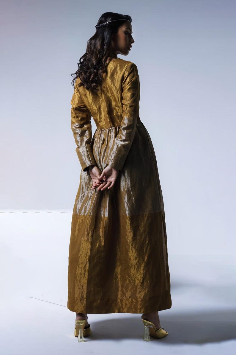 May include: A long, golden-brown dress with long sleeves and a high collar. The dress has a flowing skirt and is paired with gold-colored heels. The model is facing away from the camera, with hands clasped behind the back.