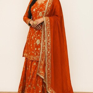May include: An orange salwar kameez, featuring detailed gold embroidery. The outfit comprises a long-sleeved tunic, flared trousers, and a matching dupatta. Gold embellishments are visible on the edges and across the fabric, giving a luxurious appearance. The outfit is suitable for special occasions.