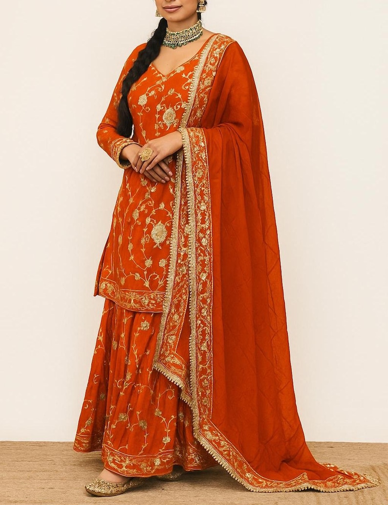 May include: An orange salwar kameez, featuring detailed gold embroidery. The outfit comprises a long-sleeved tunic, flared trousers, and a matching dupatta. Gold embellishments are visible on the edges and across the fabric, giving a luxurious appearance. The outfit is suitable for special occasions.