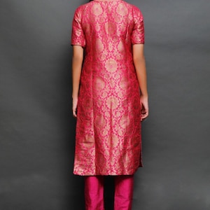 May include: A knee-length tunic with short sleeves in a vibrant fuchsia pink featuring a repeating gold floral brocade pattern, paired with matching solid fuchsia pink straight-leg trousers. The ensemble is shown from the back view.