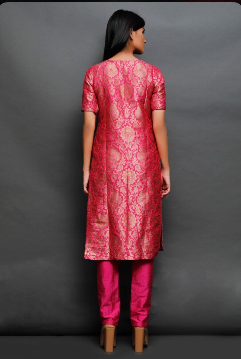 May include: A knee-length tunic with short sleeves in a vibrant fuchsia pink featuring a repeating gold floral brocade pattern, paired with matching solid fuchsia pink straight-leg trousers. The ensemble is shown from the back view.