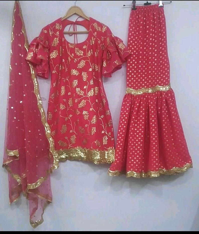 Peut inclure: Un ensemble trois pi&egrave;ces rose vif compos&eacute; d'une tunique &agrave; manches &agrave; volants avec des motifs floraux dor&eacute;s, d'une jupe &eacute;vas&eacute;e assortie &agrave; ourlet dor&eacute; et d'une dupatta rose transparente avec des paillettes dor&eacute;es et une bordure festonn&eacute;e.