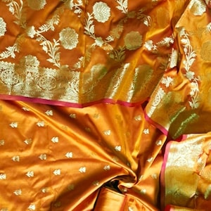 May include: A golden yellow silk saree with a floral pattern in gold and silver thread. The saree has a pink and gold border.