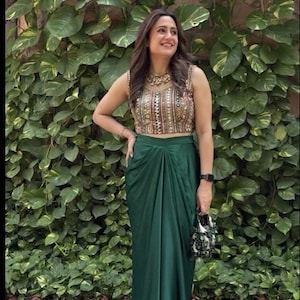 May include: A sleeveless, embellished crop top and a green draped skirt. The top has intricate beadwork and embroidery. The skirt has a high waist and a flowing, asymmetrical design. The outfit is paired with embellished heels and a small handbag.
