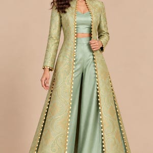May include: A sage green three-piece ensemble featuring a sleeveless crop top, high-waisted wide-leg trousers, and a long-sleeved floor-length duster coat. The coat is crafted from a patterned fabric and features gold coin-style trim along the edges.