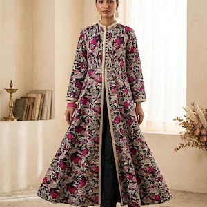 Pu&ograve; includere: Lungo cappotto aperto sul davanti con sfondo nero e un vivace motivo floreale nei toni del rosa e del bianco. Il cappotto presenta un colletto alla coreana e finiture dorate. La modella indossa pantaloni neri e scarpe dorate.