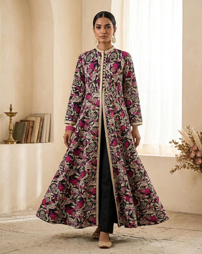 Pu&ograve; includere: Lungo cappotto aperto sul davanti con sfondo nero e un vivace motivo floreale nei toni del rosa e del bianco. Il cappotto presenta un colletto alla coreana e finiture dorate. La modella indossa pantaloni neri e scarpe dorate.
