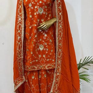 May include: An orange embroidered outfit, comprising a top, trousers, and a shawl, is displayed on a mannequin. The outfit features intricate gold floral embroidery. The shawl and edges of the outfit are trimmed with a gold border. A green beaded necklace is worn.