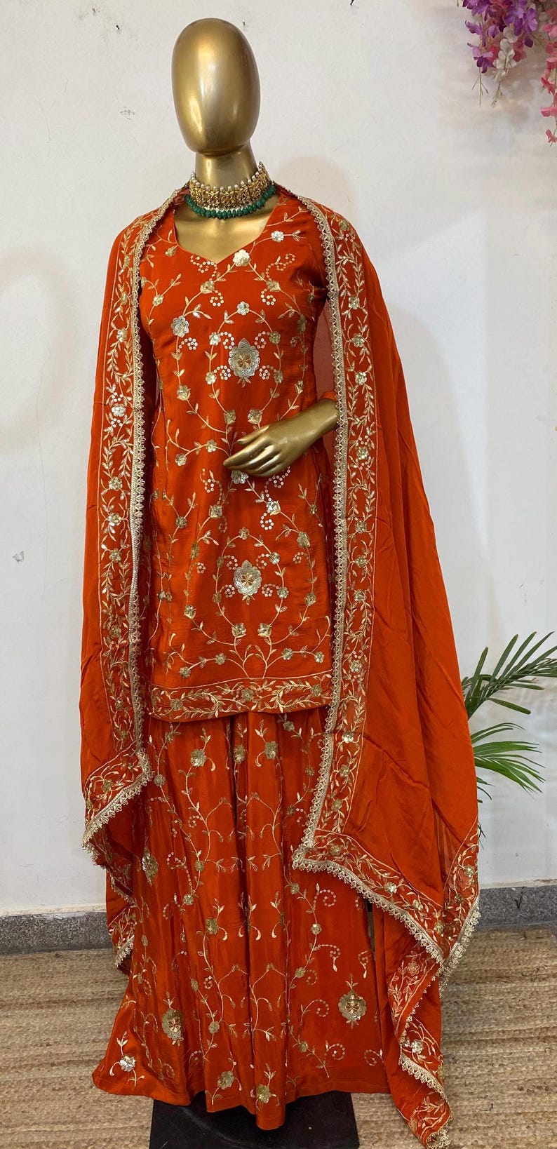 May include: An orange embroidered outfit, comprising a top, trousers, and a shawl, is displayed on a mannequin. The outfit features intricate gold floral embroidery. The shawl and edges of the outfit are trimmed with a gold border. A green beaded necklace is worn.