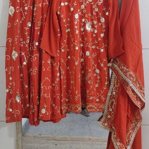 May include: A three-piece, rust-coloured outfit featuring a long-sleeved tunic, matching skirt, and a scarf. The ensemble is adorned with gold floral embroidery and sequins. The skirt and scarf have a gold trim.