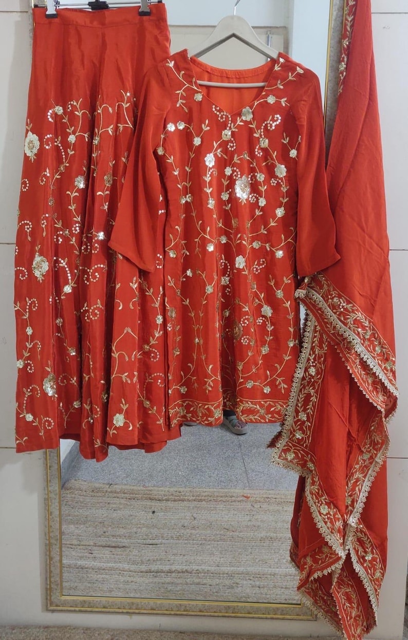 May include: A three-piece, rust-coloured outfit featuring a long-sleeved tunic, matching skirt, and a scarf. The ensemble is adorned with gold floral embroidery and sequins. The skirt and scarf have a gold trim.