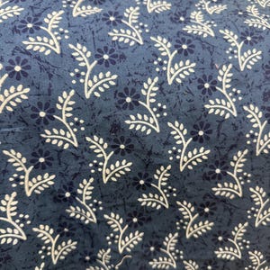 May include: A blue fabric featuring a repeating pattern of small dark blue flowers and white leafy sprigs. The background has a subtle, mottled texture in varying shades of blue, creating a layered appearance across the entire surface of the textile.