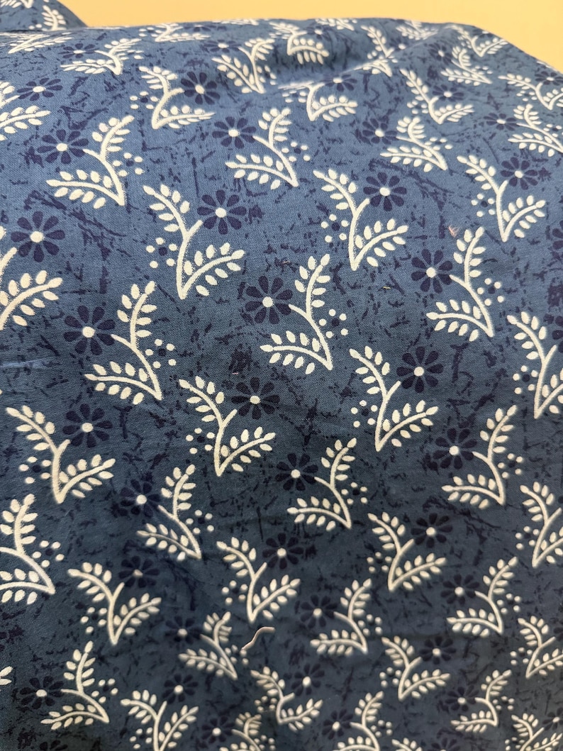 May include: A blue fabric featuring a repeating pattern of small dark blue flowers and white leafy sprigs. The background has a subtle, mottled texture in varying shades of blue, creating a layered appearance across the entire surface of the textile.