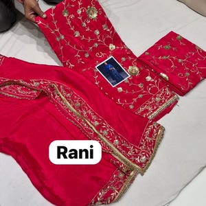 May include: A bright red salwar kameez set featuring detailed gold embroidery. The set comprises a top, trousers, and a scarf, all decorated with floral and leaf designs. The word "Rani" is displayed on the bottom of the image.