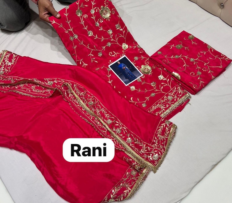 May include: A bright red salwar kameez set featuring detailed gold embroidery. The set comprises a top, trousers, and a scarf, all decorated with floral and leaf designs. The word "Rani" is displayed on the bottom of the image.