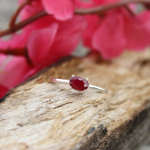 May include: A silver ring featuring a vibrant, oval-cut ruby gemstone. The ring is set against a rustic wooden surface, with bright pink flowers in the background, creating a contrast of textures and colors. A piece of jewelry.