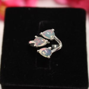 Peut inclure: Bague en argent ornée de trois opales en forme de poire. Les opales présentent un éclat irisé, affichant un éventail de couleurs. La bague est posée sur un fond de velours noir, avec des fleurs roses en arrière-plan.