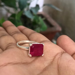 May include: A silver ring featuring a large, square-cut, deep red gemstone. The ring's band is a simple, polished silver, and the gemstone is set in a square bezel. The ring is displayed on a hand, with a blurred green background.