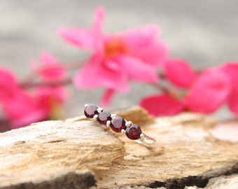 Può includere: Un anello d'argento con cinque pietre preziose rotonde, rosso scuro, disposte in fila. L'anello è appoggiato su un pezzo di legno, con fiori rosa acceso sullo sfondo sfocato. Un gioiello per un'occasione speciale.