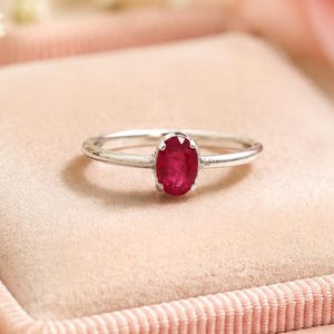 Könnte beinhalten: Ein Silberring mit einem einzelnen oval geschliffenen roten Rubin in einer Krappenfassung. Der Ring liegt auf einem weichen, hellrosa Samtkissen und präsentiert die polierte Metallschiene sowie die leuchtend rote Farbe des Edelsteins.