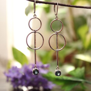 May include: Silver-toned dangle earrings with a modern design. Each earring features two connected circles, a vertical bar, and a small, round, teal-colored gemstone at the bottom. The earrings are displayed against a blurred background of green foliage and purple flowers.