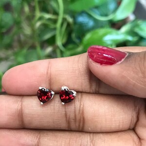 May include: A pair of heart-shaped stud earrings with a vibrant red gemstone set in a silver metal. The earrings are held in a hand, with a blurred green background.