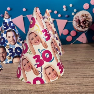 Op de afbeelding: Een set kleurrijke feestmutsen met een witte achtergrond en een herhalend patroon van gezichten en het getal 30. De mutsen zijn gemaakt van papier en hebben een kegelvorm.