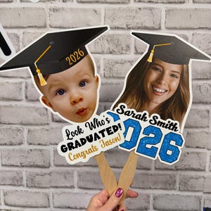 May include: Graduation photo props featuring a baby and a young woman, each wearing a graduation cap. The props include the year "2026" and the text "Look Who's GRADUATED! Congrats Jason!" and "Sarah Smith 026".