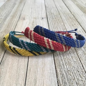 May include: Three friendship bracelets in yellow, green, and blue with black, white, and red stripes. The bracelets are woven together and are on a wooden surface.
