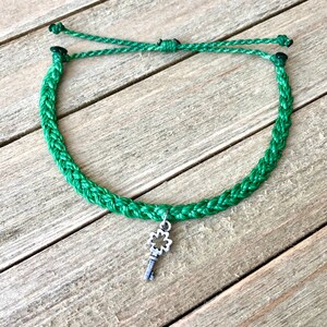 Puede incluir: Un brazalete de cordón trenzado verde con un dije de llave plateado con diseño de trébol de cuatro hojas.